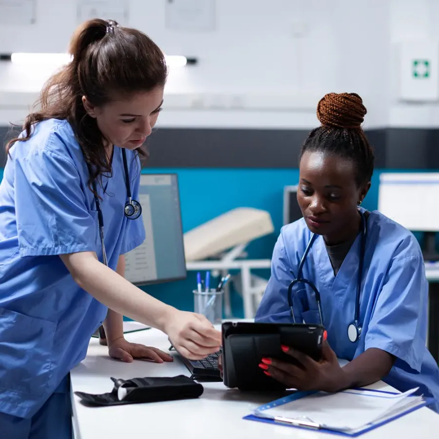 Two medical assistants in blue scrubs viewing patient charts on tablet